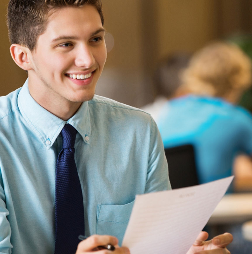 Intern in blue shirt and tie holding a document during a business meeting, seated across from another person at a table; background includes others at tables with blurred faces for privacy.