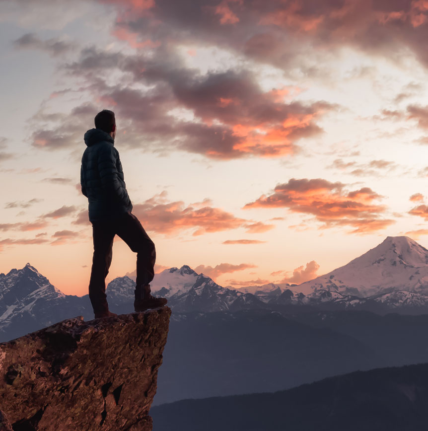 Person standing on cliff looking at sunrise over mountains