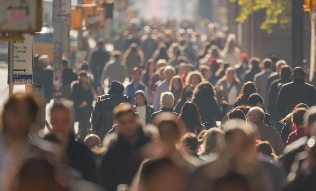 A crowded city street with people walking in various directions under bright sunlight.