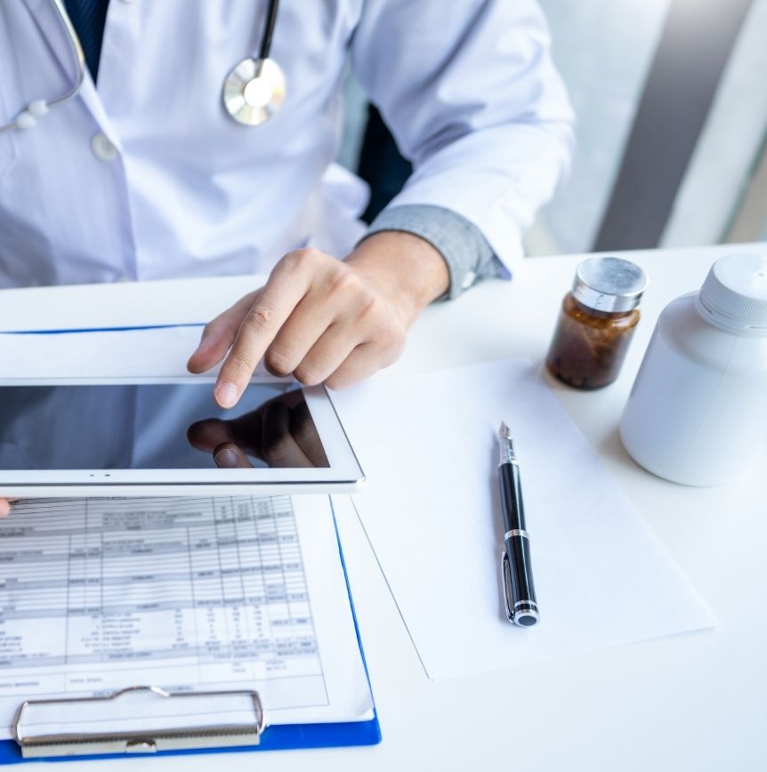 Doctor in white coat sitting at a desk with medical documents and containers using a tablet