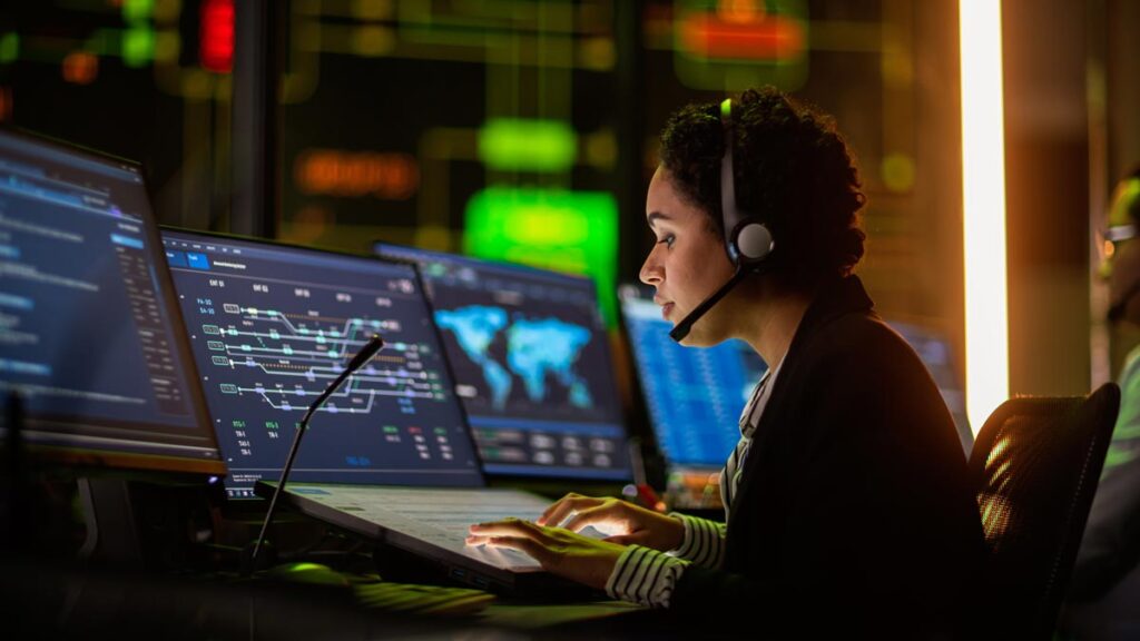 A government worker at a control center surrounded by computer screens showing data and maps.