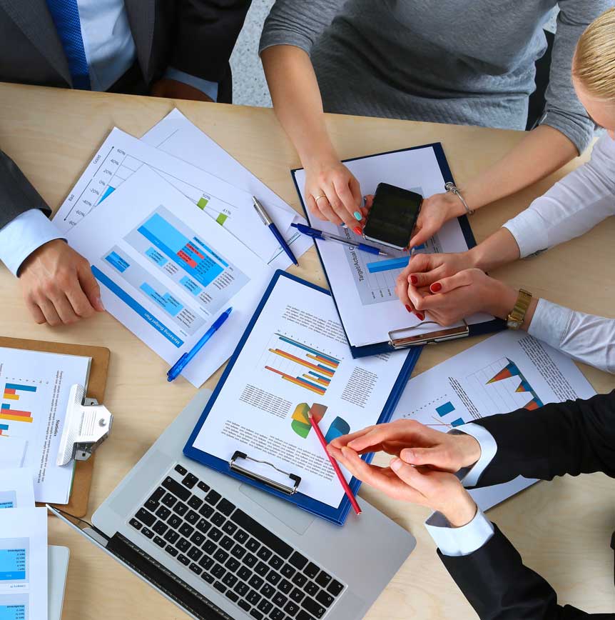 Team collaborating at a table with charts, graphs, documents, and a laptop; hands holding pens and a phone.