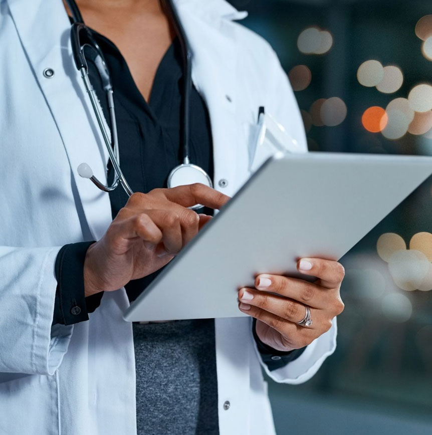 Doctor in white coat using a tablet, stethoscope visible, with blurred lights in the background.