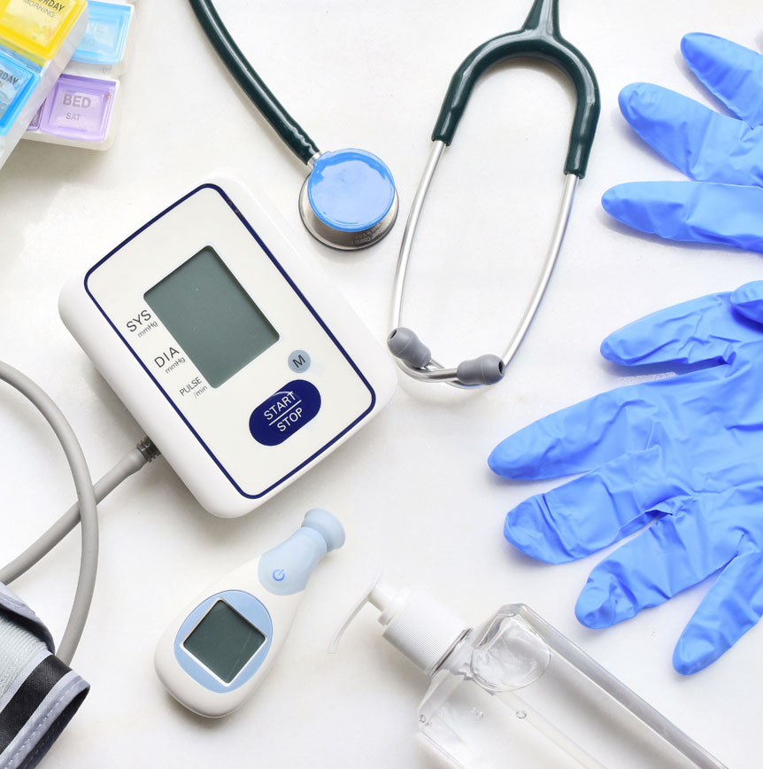 Medical equipment arranged on a white surface, including a digital blood pressure monitor, stethoscope, blue disposable gloves, thermometer, pill organizer, and a clear pump bottle.