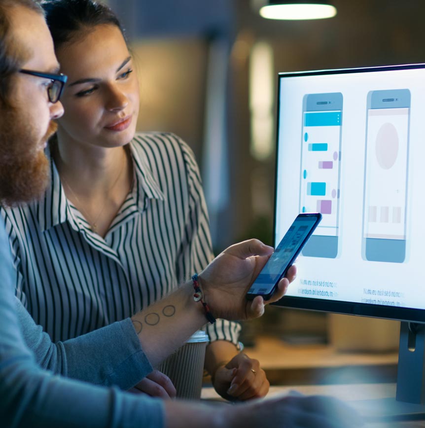 Two people reviewing a mobile app design on a computer screen while one demonstrates features on a smartphone.