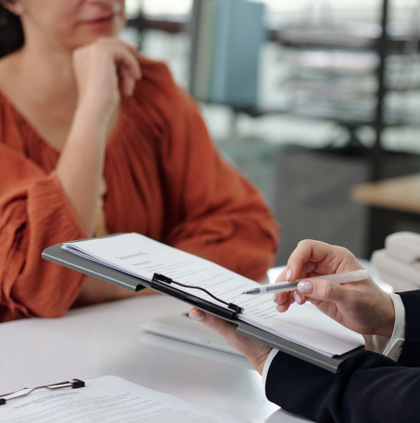 Co-workers reviewing documents on a clipboard, with one person pointing to a form using a pen during a professional consultation.