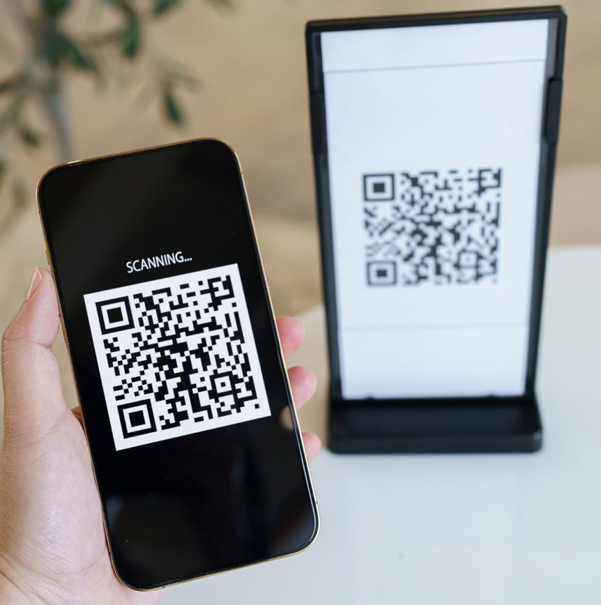 Close-up of a hand holding a smartphone scanning a QR code labeled “SCANNING…” while pointed at a matching QR code displayed on a stand in the background