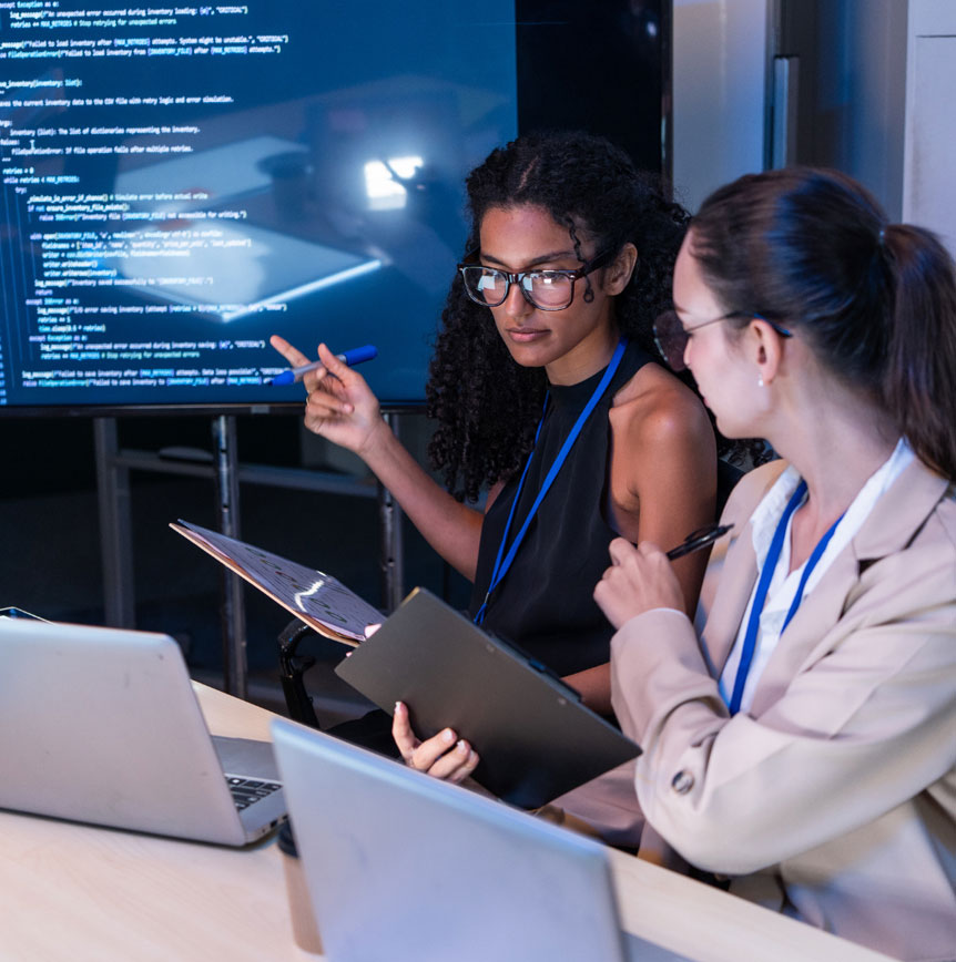 Two people review code on a large screen while working with laptops and clipboards in a collaborative technology setting.