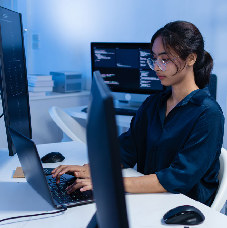 A person working on a laptop at a desk with multiple computer monitors displaying code in an office environment, illustrating practical AI and software development work