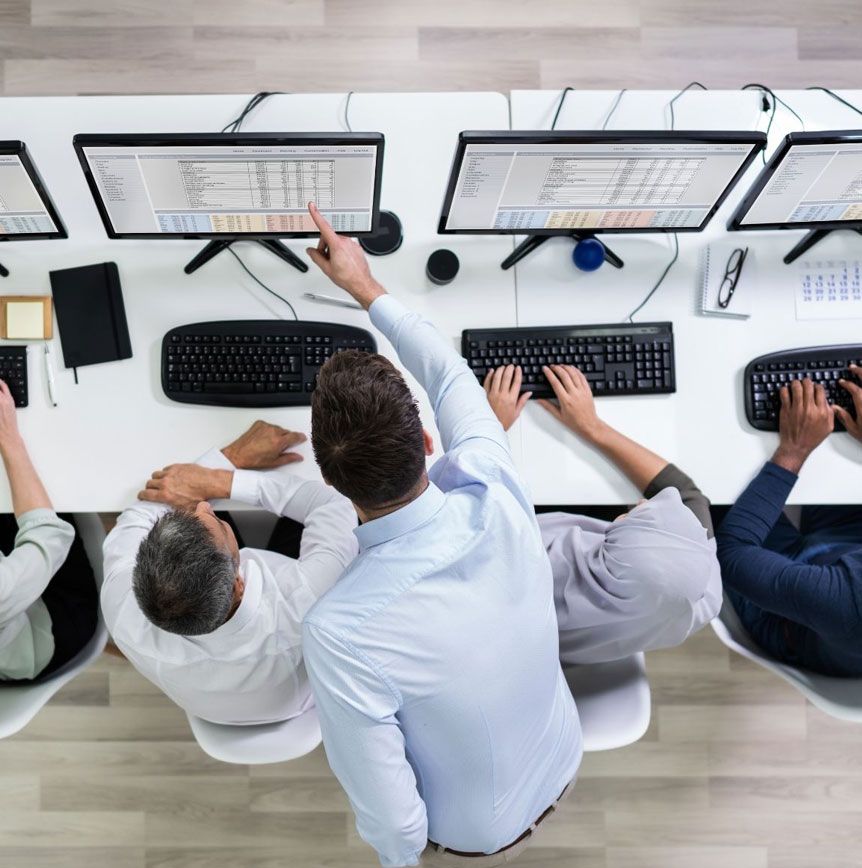 Overhead view of a team working at a row of computer workstations, with one person pointing to data displayed on a monitor while others type on keyboards at a shared desk.
