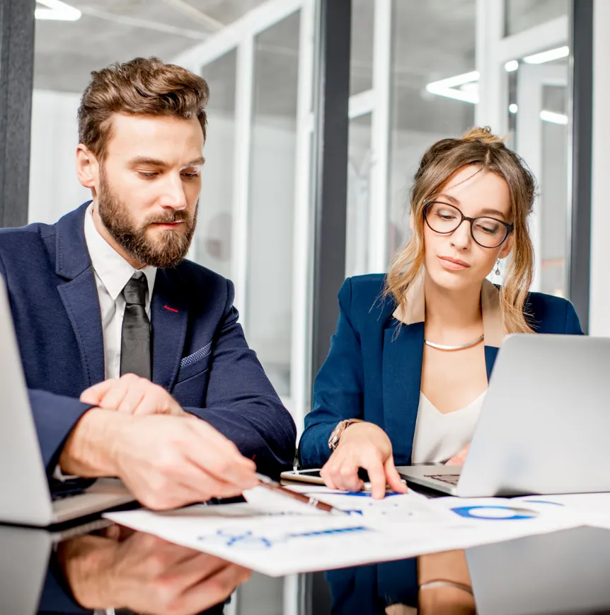 A bearded man in a navy suit points a pen at printed reports with blue charts while a woman in glasses and a navy blazer examines them beside her laptop, at a glossy desk in a bright glass-walled office.