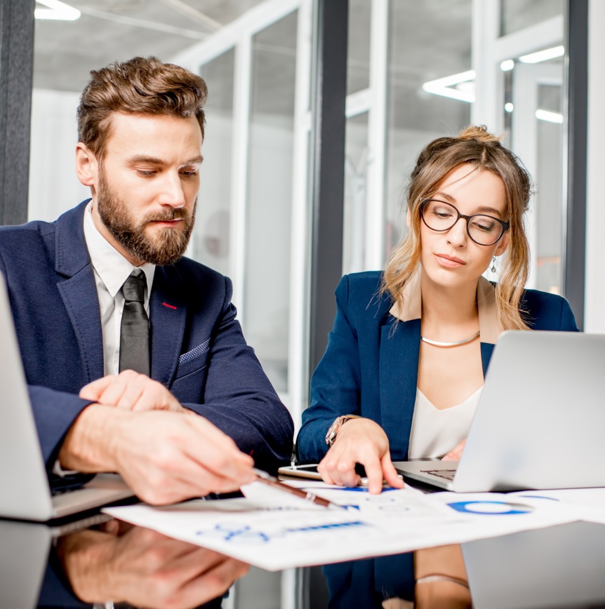Two business professionals review printed charts and documents at a conference table while working on laptop computers in a modern office setting.