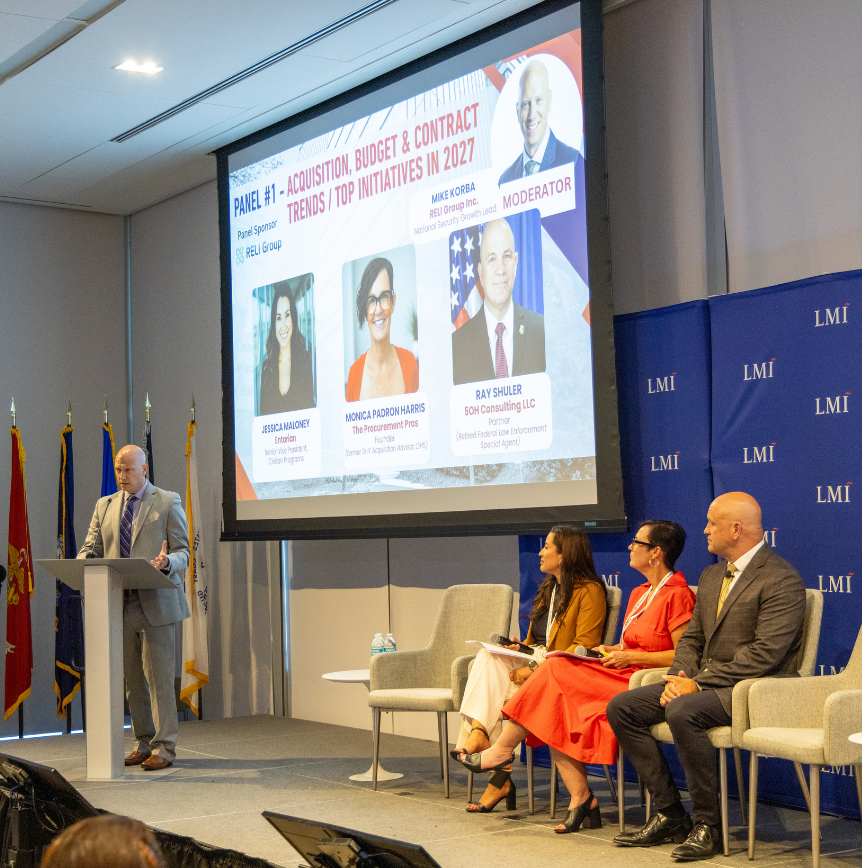 RELI Group's Mike Korba at a lectern beside three seated panelists during a discussion on acquisition, budget and contract trends for DHS initiatives in 2027.